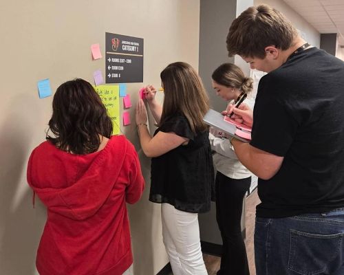  Students stand at wall with postits and pencils to work on a grammar problem